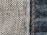 Close-up view of denim fabric showcasing a detailed weave pattern in blue and white. A seam with visible stitches runs vertically on the right side, highlighting the texture and craftsmanship.
