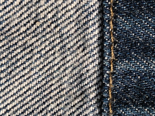Close-up view of denim fabric showcasing a detailed weave pattern in blue and white. A seam with visible stitches runs vertically on the right side, highlighting the texture and craftsmanship.
