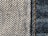 Close-up view of denim fabric showcasing a detailed weave pattern in blue and white. A seam with visible stitches runs vertically on the right side, highlighting the texture and craftsmanship.