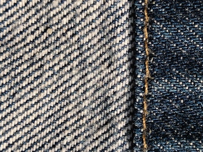 Close-up view of denim fabric showcasing a detailed weave pattern in blue and white. A seam with visible stitches runs vertically on the right side, highlighting the texture and craftsmanship.