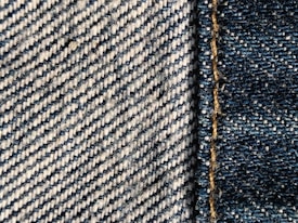 Close-up view of denim fabric showcasing a detailed weave pattern in blue and white. A seam with visible stitches runs vertically on the right side, highlighting the texture and craftsmanship.