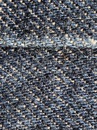 A close-up view of a textured fabric with interwoven dark blue and beige threads, creating a detailed woven pattern. The fibers appear slightly fuzzy, indicating a tightly woven, rough textile.