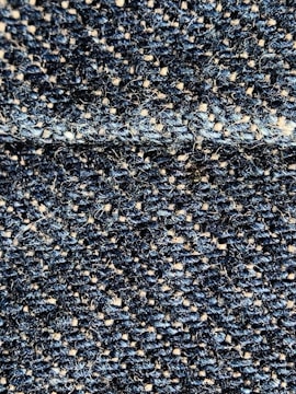 A close-up view of a textured fabric with interwoven dark blue and beige threads, creating a detailed woven pattern. The fibers appear slightly fuzzy, indicating a tightly woven, rough textile.