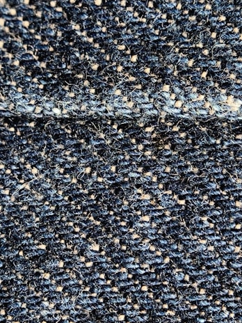 A close-up view of a textured fabric with interwoven dark blue and beige threads, creating a detailed woven pattern. The fibers appear slightly fuzzy, indicating a tightly woven, rough textile.