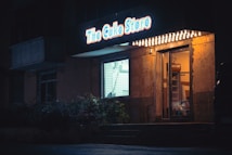 A bakery storefront is dimly lit at night. The bright sign above the door displays the name 'The Cake Store' in glowing blue and red colors. Warm light spills out from the interior through the glass entrance and window, casting a cozy atmosphere. The exterior appears aged with a rustic look, surrounded by shadows and foliage.