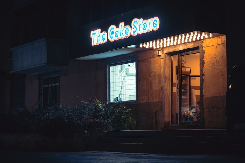 A bakery storefront is dimly lit at night. The bright sign above the door displays the name 'The Cake Store' in glowing blue and red colors. Warm light spills out from the interior through the glass entrance and window, casting a cozy atmosphere. The exterior appears aged with a rustic look, surrounded by shadows and foliage.