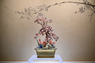 A decorative floral arrangement with pink and red blossoms is elegantly displayed in an ornate golden pot. The background features a beige wall with subtle black line art depicting branches, which complements the delicate design of the flower piece.