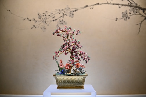 A decorative floral arrangement with pink and red blossoms is elegantly displayed in an ornate golden pot. The background features a beige wall with subtle black line art depicting branches, which complements the delicate design of the flower piece.