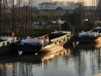 Three large cargo barges with flags are docked closely together on a calm river. The scene is set during a sunlit period, causing warm reflections on the water's surface. In the background, there are tall bare trees and a mix of industrial and residential buildings, indicating an urban setting.