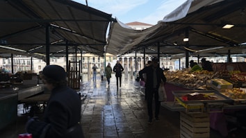 A covered outdoor market with multiple vendors displaying fresh produce and various goods on stalls. People are walking through the market, including some carrying bags with purchases. The scene is set under a canopy, with the sunlight casting shadows on the wet cobblestone ground.