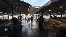 A covered outdoor market with multiple vendors displaying fresh produce and various goods on stalls. People are walking through the market, including some carrying bags with purchases. The scene is set under a canopy, with the sunlight casting shadows on the wet cobblestone ground.