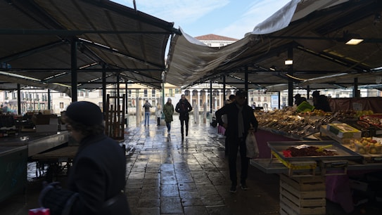 A covered outdoor market with multiple vendors displaying fresh produce and various goods on stalls. People are walking through the market, including some carrying bags with purchases. The scene is set under a canopy, with the sunlight casting shadows on the wet cobblestone ground.