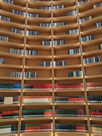 Rows of books neatly arranged on curved wooden bookshelves, with lighting that accentuates the book spines. The bookshelves are filled with a variety of book colors, mainly red, green, and blue, creating a visually appealing pattern.