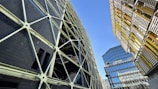 A complex urban landscape featuring modern architecture with geometric designs. A striking dark building with a crisscross pattern of supports contrasts against another structure with yellow and metallic horizontal slats. In the background, a reflective glass building rises, adding to the urban feel. The sky is clear and blue, providing a stark contrast to the man-made structures.