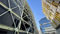 A complex urban landscape featuring modern architecture with geometric designs. A striking dark building with a crisscross pattern of supports contrasts against another structure with yellow and metallic horizontal slats. In the background, a reflective glass building rises, adding to the urban feel. The sky is clear and blue, providing a stark contrast to the man-made structures.