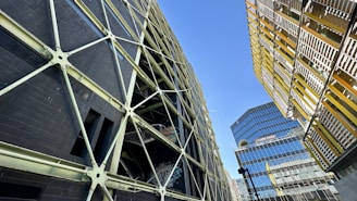 A complex urban landscape featuring modern architecture with geometric designs. A striking dark building with a crisscross pattern of supports contrasts against another structure with yellow and metallic horizontal slats. In the background, a reflective glass building rises, adding to the urban feel. The sky is clear and blue, providing a stark contrast to the man-made structures.