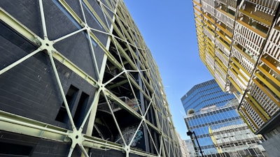 A complex urban landscape featuring modern architecture with geometric designs. A striking dark building with a crisscross pattern of supports contrasts against another structure with yellow and metallic horizontal slats. In the background, a reflective glass building rises, adding to the urban feel. The sky is clear and blue, providing a stark contrast to the man-made structures.