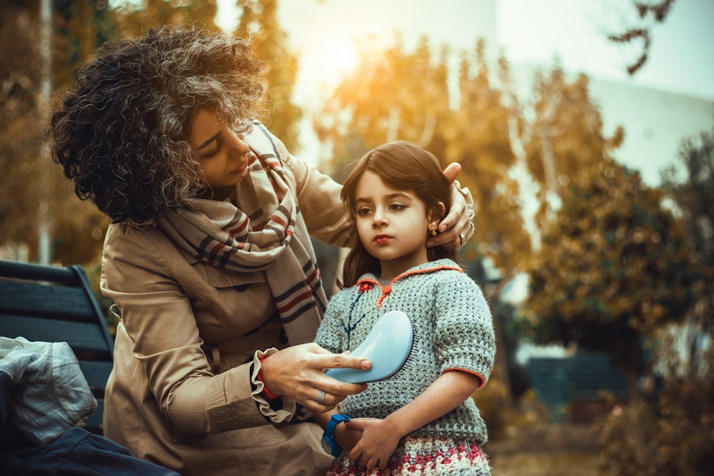 a woman holding a young girl's hair while sitting on a bench