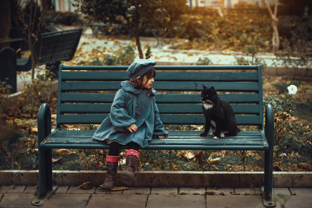 a little girl sitting on a bench next to a black cat