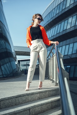 A stylishly dressed woman stands confidently on a set of stone stairs between two modern glass buildings. She wears a bright orange jacket over a black top, complemented by beige pants and high heels. The sun is shining, creating a strong contrast with the architectural background.