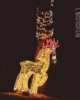 A festive outdoor setup featuring a large, illuminated reindeer made of golden lights stands beside a tree. The tree is wrapped in spiraling strings of white lights. Above the reindeer, a decorative element shaped like antlers is lit with red lights, and a figure resembling Santa or another festive character is also made of lights.