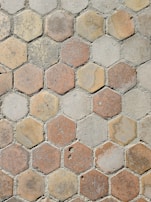 A pattern of hexagonal tiles with a weathered appearance, featuring shades of beige, brown, and gray. The tiles have varying amounts of wear and texture, giving a rustic and aged look. The grout between the tiles is visible and uneven, suggesting an outdoor pavement or walkway.