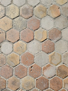 A pattern of hexagonal tiles with a weathered appearance, featuring shades of beige, brown, and gray. The tiles have varying amounts of wear and texture, giving a rustic and aged look. The grout between the tiles is visible and uneven, suggesting an outdoor pavement or walkway.