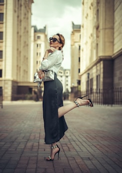 A stylish woman is standing on one leg in an urban environment. She wears sunglasses, a white blouse, and a dark, flowing skirt with a high slit, paired with high-heeled sandals decorated with jewels. She holds a silver clutch in her hand, leaning slightly to one side in a confident pose between two tall buildings with classical architecture.