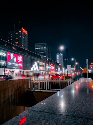 A bustling cityscape at night with towering buildings illuminated by bright lights and billboards. Cars are seen moving along the street, creating a lively atmosphere. Several advertisements and store signs are visible, including a prominent Adidas logo and a 'Half Price' sale sign. The scene is vibrant, with a significant presence of commercial activity.
