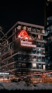 A nighttime urban scene featuring a modern building adorned with bright, festive lights. A neon sign with a red Santa hat and the words 'Browary Warszawskie' stands out against the dark sky. Snow is visible on the ground, suggesting a winter setting, and the building's windows are illuminated.