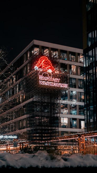 A nighttime urban scene featuring a modern building adorned with bright, festive lights. A neon sign with a red Santa hat and the words 'Browary Warszawskie' stands out against the dark sky. Snow is visible on the ground, suggesting a winter setting, and the building's windows are illuminated.