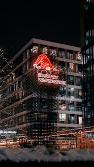 A nighttime urban scene featuring a modern building adorned with bright, festive lights. A neon sign with a red Santa hat and the words 'Browary Warszawskie' stands out against the dark sky. Snow is visible on the ground, suggesting a winter setting, and the building's windows are illuminated.