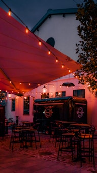 An outdoor seating area with string lights hanging above, casting a warm glow. Several black tables and stools are placed on a cobblestone pavement. There is a black food truck parked nearby with a sign saying 'The Pizza Box'. The scene is set against a white building, partially covered by a red canopy.