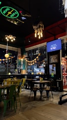 The interior of a casual restaurant or cafe with modern decor and a cozy atmosphere. Several wooden tables and chairs are neatly arranged, and a mix of green and yellow chairs provides a vibrant touch. Decorative lights hang from the ceiling, and pumpkins or similar decorations are strung across the window. A sign promotes 'Back to the Show' on a screen, and part of a neon sign is visible.