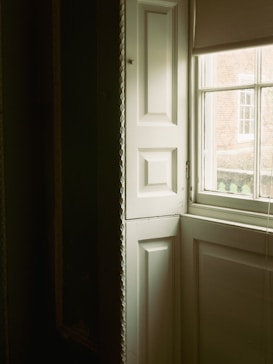 A partially open window with white, paneled shutters allowing soft natural light to stream into a room. The view through the window shows a red brick building in the background. The interior wall is dark, creating a contrasting ambiance with the light.