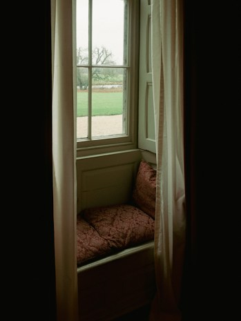 A cozy window nook with a view of an outdoor landscape. The nook features plush, patterned cushions and is framed by soft, flowing curtains. Outside, a sprawling green lawn and a tree are visible, providing a serene and peaceful atmosphere.