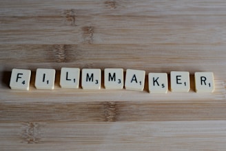 Scrabble tiles arranged to spell the word 'FILMMAKER' placed on a wooden surface. Each tile displays a letter and a corresponding numeric value.