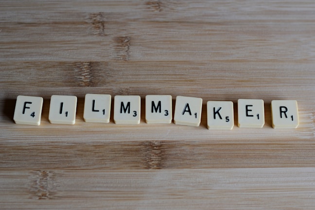Scrabble tiles arranged to spell the word 'FILMMAKER' placed on a wooden surface. Each tile displays a letter and a corresponding numeric value.