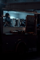 A dimly lit food truck with two people inside, one on the phone and the other interacting with large metal containers. A sign advertising mulled wine is visible next to them. The setting conveys a nighttime scene with a cozy, intimate ambiance.