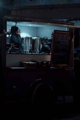 A dimly lit food truck with two people inside, one on the phone and the other interacting with large metal containers. A sign advertising mulled wine is visible next to them. The setting conveys a nighttime scene with a cozy, intimate ambiance.