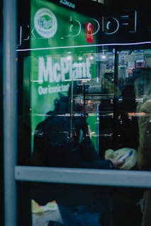 Reflections on a glass pane display part of a fast food establishment with signage promoting a 'McPlant' product. The interior shows shelves with various items, and there are blurry figures both inside and outside holding mobile devices.