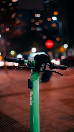 A close-up view of the handlebars of a green electric scooter with the brand name 'Bolt' visible on the handlebar stem. The background is a cityscape at night with blurred lights and traffic, creating a bokeh effect.