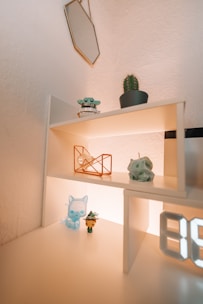 A minimalist shelf setup featuring small toys and decorative objects. A small cactus in a pot is placed on the top shelf next to a robotic figure. Below, on the middle shelf, there is a geometric wireframe sculpture and a small figurine of a character with a long snout. On the bottom shelf, there's a translucent blue cat-like figure and a character wearing a green outfit with a feathered hat. An illuminated digital number display is partially visible.