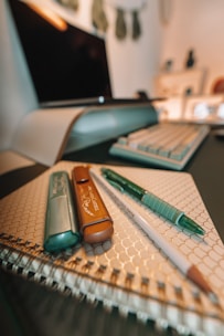 A neatly arranged set of writing tools including a highlighter, a pencil, a marker, and a pen placed on top of a spiral-bound notebook with a geometric patterned cover. In the background, a blurred computer monitor and keyboard can be seen, suggesting a workspace environment.