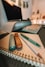A neatly arranged set of writing tools including a highlighter, a pencil, a marker, and a pen placed on top of a spiral-bound notebook with a geometric patterned cover. In the background, a blurred computer monitor and keyboard can be seen, suggesting a workspace environment.