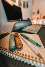 A neatly arranged set of writing tools including a highlighter, a pencil, a marker, and a pen placed on top of a spiral-bound notebook with a geometric patterned cover. In the background, a blurred computer monitor and keyboard can be seen, suggesting a workspace environment.