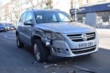 A silver Volkswagen SUV with noticeable front-end damage parked on a city street. The vehicle's front bumper and headlight are significantly dented and scratched. There are small debris pieces scattered around the tires, suggesting a recent collision. Surrounding the car are other parked vehicles and a building with a closed store featuring a 'For Sale' sign.