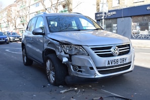 A silver Volkswagen SUV with noticeable front-end damage parked on a city street. The vehicle's front bumper and headlight are significantly dented and scratched. There are small debris pieces scattered around the tires, suggesting a recent collision. Surrounding the car are other parked vehicles and a building with a closed store featuring a 'For Sale' sign.