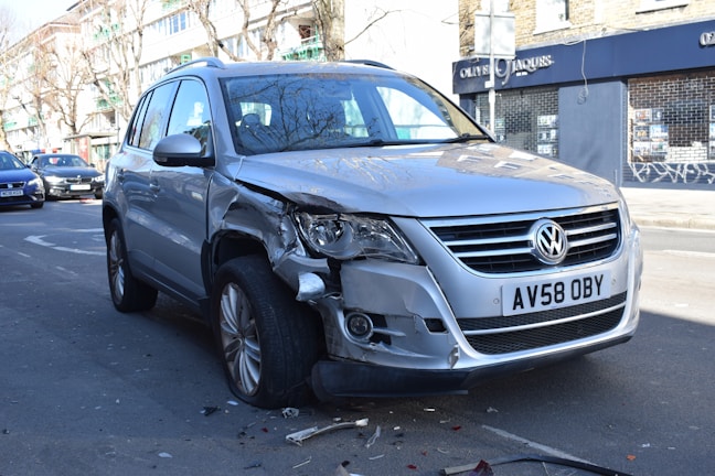 A silver Volkswagen SUV with noticeable front-end damage parked on a city street. The vehicle's front bumper and headlight are significantly dented and scratched. There are small debris pieces scattered around the tires, suggesting a recent collision. Surrounding the car are other parked vehicles and a building with a closed store featuring a 'For Sale' sign.