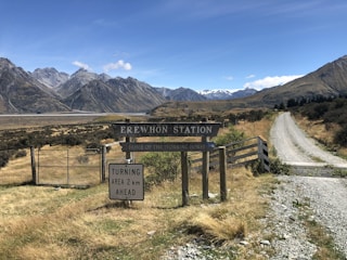A rural landscape featuring a gravel road leading towards a mountainous horizon. The foreground has a sign reading 'Erewhon Station' and 'Home of the Working Horse', and a directional sign indicating a turning area 2 km ahead. The area is surrounded by dry grasslands with tall mountains in the background under a clear blue sky.